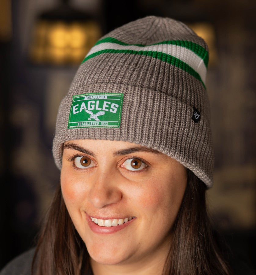 A woman wears the penobscot cuff knit beanie. It's grey with a green patch on the front.