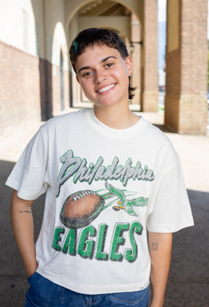 A smiling person standing outdoors in front of a brick building, wearing a white graphic T-shirt with 'Philadelphia Eagles' text and a football illustration, paired with blue jeans.