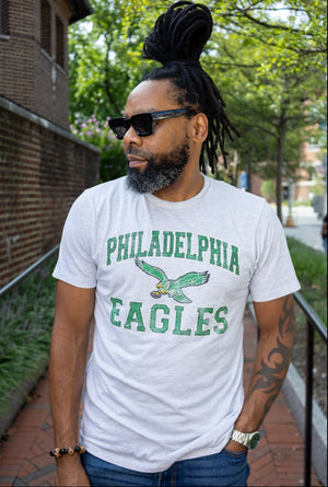 A man wearing a white graphic t-shirt with 'PHILADELPHIA EAGLES' and an eagle logo printed in green, paired with blue jeans and black sunglasses, standing outdoors on a brick walkway.