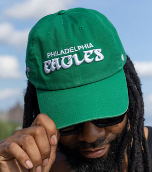 A man wearing a green Philadelphia Eagles baseball cap, sunglasses, and a black tank top, holding the brim of the cap with his hand against an outdoor sky background.