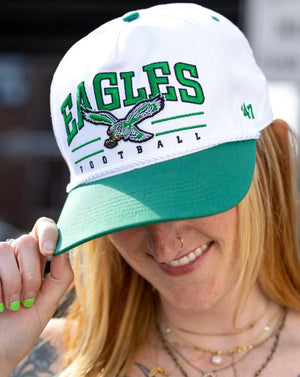 A woman wearing a white and green Eagles Football trucker hat, holding the brim with her hand. The hat features green lettering, a green and white eagle graphic, and a green '47' logo on the side.